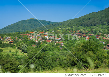 Traditional Bulgarian village with red rooftops nestled in green hills under clear blue sky. Ethnographic landscape blends eco-lifestyle, vernacular architecture and rural charm 125039745