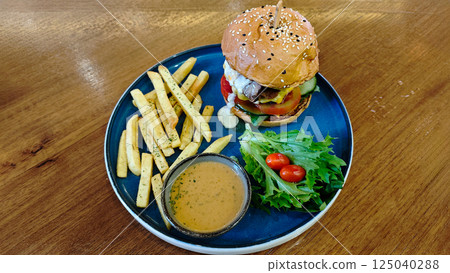 Delicious Gourmet Cheeseburger with Fries, Salad, and Dipping Sauce on a Blue Plate in a Rustic Wooden Table Setting, Featuring Fresh Ingredients and Tasty Sides 125040288