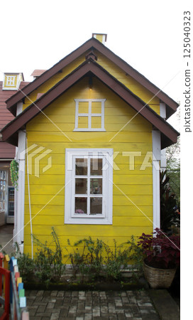 Charming Yellow Wooden House Facade with White Trim and Rustic Window, Surrounded by Lush Greenery in a Quaint Village Setting 125040323