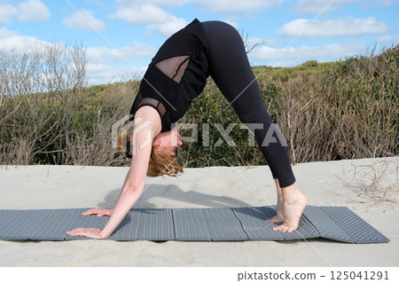 Woman practicing yoga outdoors on sandy surface, performing downward dog pose on a mat, surrounded by nature under blue sky with light clouds, peaceful atmosphere. 125041291