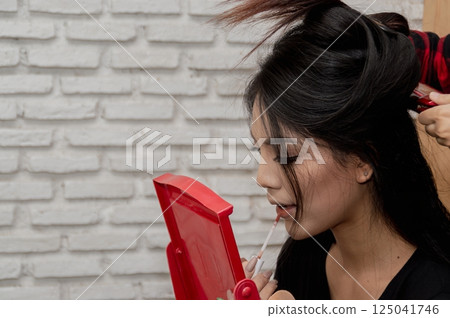 A young woman styling her hair while looking in a handheld mirror, showcasing personal grooming and beauty routine. A young woman styling her hair while looking in a handheld mirror, showcasing personal grooming and beauty routine. 125041746
