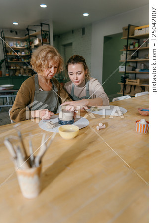 Teacher guides an elderly women, demonstrating sculpting techniques in a hands-on class. Elderly Caucasian woman learns pottery at a creative workshop with a young mentor 125042007