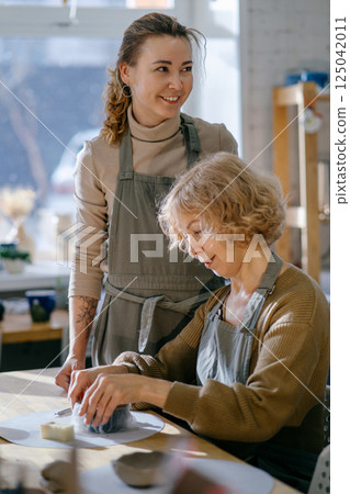 Teacher guides an elderly women, demonstrating sculpting techniques in a hands-on class. Elderly Caucasian woman learns pottery at a creative workshop with a young mentor Teacher guides an elderly women, demonstrating sculpting techniques in a hands-on class. Elderly Caucasian woman learns pottery at a creative workshop with a young mentor 125042011