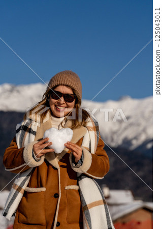 Snow Heart Woman Mountains Winter: Joyful woman holds snow heart, sunny winter day in snowy mountains. 125043011