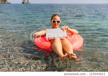 A woman is sitting on a pink inflatable raft in the ocean, using a laptop. Concept of relaxation and leisure, as the woman is enjoying her time in the water while working on her laptop. A woman is sitting on a pink inflatable raft in the ocean, using a laptop. Concept of relaxation and leisure, as the woman is enjoying her time in the water while working on her laptop. 125043021
