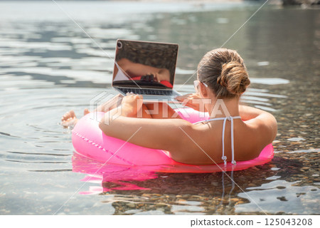 A woman is sitting on a pink inflatable raft in the water, using a laptop. Concept of relaxation and leisure, as the woman is enjoying her time in the water while working on her laptop. 125043208