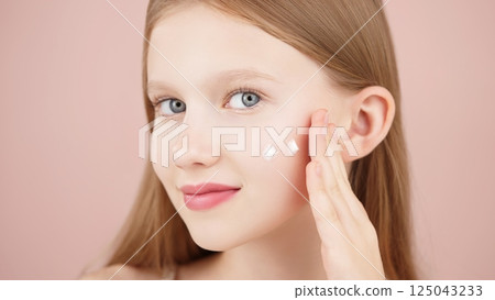 Portrait of a smiling cheerful girl applying face cream on cheek for skin glow. Advertising face cream and skincare products for teens. Blonde girl with blue eyes isolated in studio on pink background 125043233