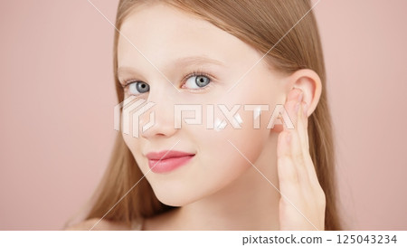 Portrait of a smiling cheerful girl applying face cream on cheek for skin glow. Advertising face cream and skincare products for teens. Blonde girl with blue eyes isolated in studio on pink background 125043234