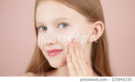 Portrait of a smiling cheerful girl applying face cream on cheek for skin glow. Advertising face cream and skincare products for teens. Blonde girl with blue eyes isolated in studio on pink background 125043235