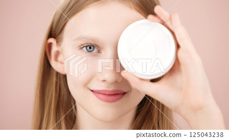 Portrait of a funny smiling blonde young girl posing in studio, holding a cosmetic cream jar in front of her eye on pink background. Advertising face cream. Skincare products for teens or kids Portrait of a funny smiling blonde young girl posing in studio, holding a cosmetic cream jar in front of her eye on pink background. Advertising face cream. Skincare products for teens or kids 125043238