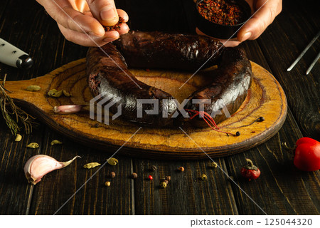Hands sprinkle spices over rich, dark sausages resting on a wooden serving board adorned with garlic, herbs, and vibrant chili. The atmosphere is warm and inviting Hands sprinkle spices over rich, dark sausages resting on a wooden serving board adorned with garlic, herbs, and vibrant chili. The atmosphere is warm and inviting 125044320