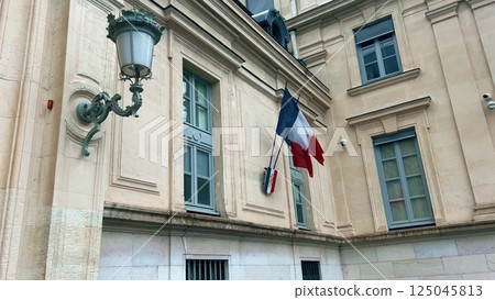 Waving national flags of France and the shield with letters RF Republique Francaise or French Republic on the typical administrative building Waving national flags of France and the shield with letters RF Republique Francaise or French Republic on the typical administrative building 125045813