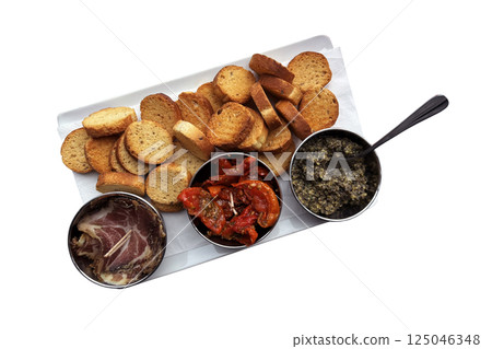 Starter dish with dried sweet tomatoes, black olive tapenade, meat slices and croutons in three bowls on rectangular plate, rustic traditional appetizer of French cuisine, restaurant menu 125046348
