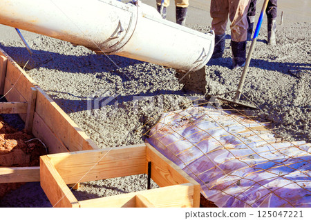 Construction workers pour wet cement from chute into wooden formwork at building site cement is being poured over rebar grid that provides structural reinforcement for concrete foundation.. 125047221