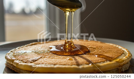 Close up of golden syrup dripping from a spoon onto warm pancakes. Morning light through window. Rich texture on pancake surface. 125050244