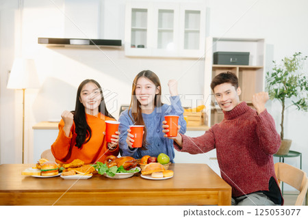 Young Asian friends enjoy a cozy dinner party at home, sharing food, drinks, and laughter in a warm modern kitchen. 125053077