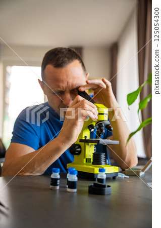 Man examining various samples under a microscope at home as a hobby. Concept of scientific curiosity and amateur microscopy 125054300