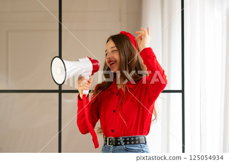 Cheerful teen girl speaking into megaphone in bright interior. Cheerful teen girl speaking into megaphone in bright interior. 125054934