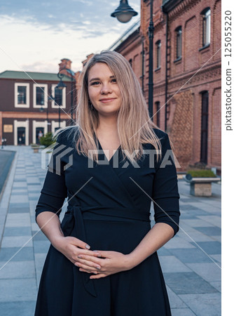 Young pregnant woman in a black dress poses confidently outside historic red brick buildings in a town square during sunset 125055220
