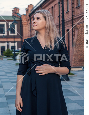 Young pregnant woman in a black dress poses confidently outside historic red brick buildings in a town square during sunset 125055223
