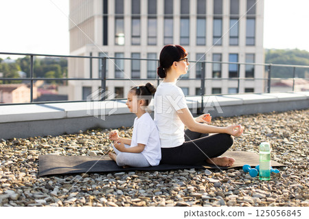 Caucasian adult woman and young girl sitting back-to-back meditating outdoors together on a rooftop. They practice yoga and mindfulness during a sunny day. Caucasian adult woman and young girl sitting back-to-back meditating outdoors together on a rooftop. They practice yoga and mindfulness during a sunny day. 125056845