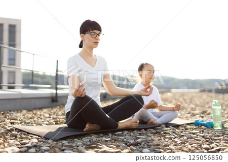 Light-skinned adult woman and young girl meditating in lotus position outdoors on mat, peaceful expressions, embracing mindfulness and calm environment. 125056850