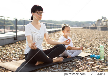 Mother and young daughter meditating together in lotus position on rooftop in morning tranquility, promoting relaxation, focus, and mindfulness. Peaceful outdoor urban setting with calming atmosphere 125056851