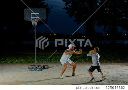 A father and son enjoy a night basketball game at a local park under bright lights focusing on teamwork and fun 125056862