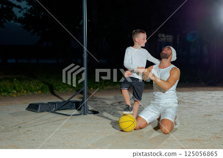 Happy family dad and son looking at camera and smiling while sitting on basketball court outdoor. Boy with handsome father after game on playground, sport concept 125056865