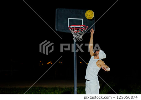 A man performs a slam dunk on a basketball court at night under the glare of bright outdoor lights 125056874