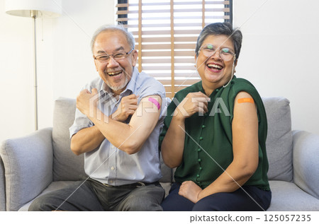 Senior Asian couple get vaccinated with bandage on arm show thumb up sign in living room. Grandfather and grandmother get vaccine. 125057235