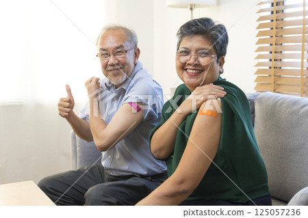Senior Asian couple get vaccinated with bandage on arm show thumb up sign in living room. Grandfather and grandmother get vaccine. Senior Asian couple get vaccinated with bandage on arm show thumb up sign in living room. Grandfather and grandmother get vaccine. 125057236