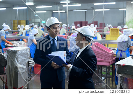 Two factory managers in safety helmets discussing documents inside a busy canned fish processing plant 125058283