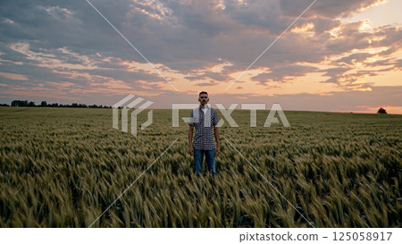 Confident farmer standing in golden wheat field, sunset casting warm light, highlighting agricultural landscape and rural prosperity 125058917