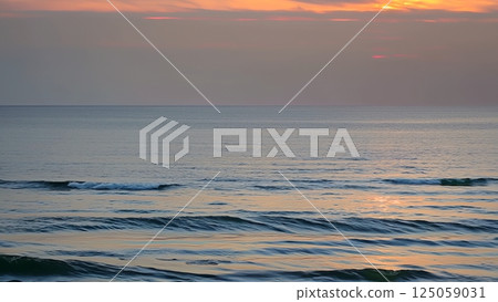 Sunset painting pink and orange sky, casting warm light over rhythmic waves gently rolling across serene ocean surface with tranquil coastal horizon 125059031