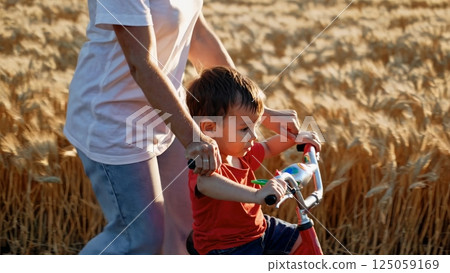 Parent supporting child learning balance bike, navigating golden wheat field with care, sharing meaningful summer moment while bonding outdoors Parent supporting child learning balance bike, navigating golden wheat field with care, sharing meaningful summer moment while bonding outdoors 125059169