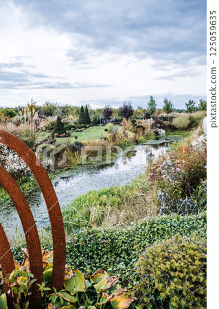 Lush landscaped garden with a reflective pond, sculpted greenery, and a rusted metal art piece in the foreground under a cloudy sky. Biodiverse planting, garden design, ecological restoration Lush landscaped garden with a reflective pond, sculpted greenery, and a rusted metal art piece in the foreground under a cloudy sky. Biodiverse planting, garden design, ecological restoration 125059635