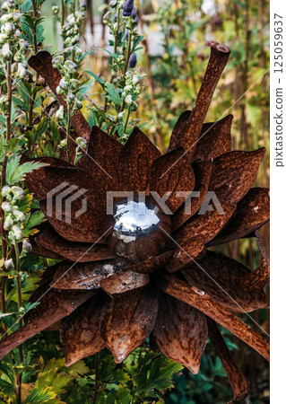 Rust-colored metal flower sculpture with a mirrored center sphere surrounded by tall garden plants. Outdoor art installation, rustic garden decor, recycled materials, nature-inspired design 125059637