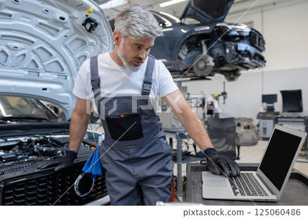 Man technician looking at inspection vehicle details under car hood using laptop for diagnostic 125060486