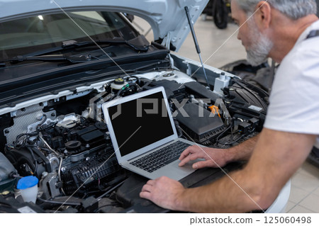 Engineer bearded man looking at inspection vehicle details under car hood using laptop Engineer bearded man looking at inspection vehicle details under car hood using laptop 125060498