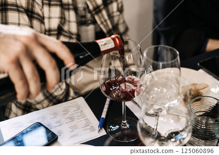 Participant pouring red wine into a glass during a wine workshop session with a tasting card nearby. Sommelier training, hands-on wine education, sensory learning, grape varietals Participant pouring red wine into a glass during a wine workshop session with a tasting card nearby. Sommelier training, hands-on wine education, sensory learning, grape varietals 125060896