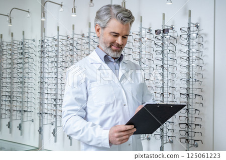 Mature doctor in lab coat standing near the stand with glasses and looking confident Mature doctor in lab coat standing near the stand with glasses and looking confident 125061223