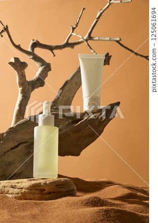 White tube without label placed on parched wooden trunk with pointed tip. Dropper bottle of serum displayed on natural stone with concave and convex edges. Presentation on a sand field in the desert. White tube without label placed on parched wooden trunk with pointed tip. Dropper bottle of serum displayed on natural stone with concave and convex edges. Presentation on a sand field in the desert. 125061694