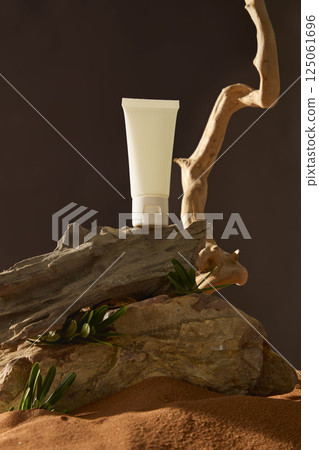 An unlabeled cream tube stands on a decayed wooden plank, its texture cracked and timeworn. The plank rests on a coarse, jagged stone, partially buried in fine desert sand. Black background. An unlabeled cream tube stands on a decayed wooden plank, its texture cracked and timeworn. The plank rests on a coarse, jagged stone, partially buried in fine desert sand. Black background. 125061696