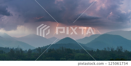 Lightning bolts strike across mountain ridges under dramatic storm clouds, capturing the power and beauty of nature during a thunderstorm over a forested landscape. 125061734