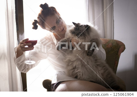Woman holding a fluffy cat and drinking red wine by the window Woman holding a fluffy cat and drinking red wine by the window 125061904