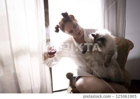 Woman drinking wine with her cat by the window 125061905