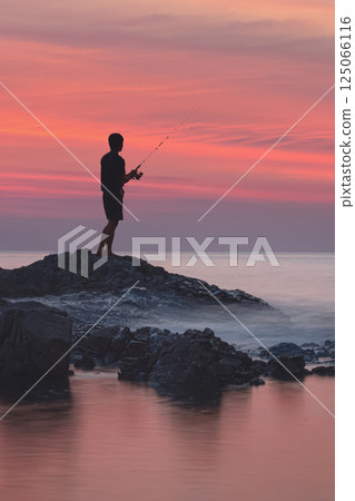 Sea fishing in the tropics. The silhouette of a fisherman on the rocks among the blurred waves and water with red highlights from the sunset. Stunning seascape 125066116