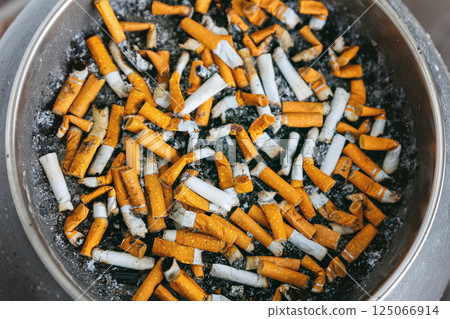 Close-up top view discarded cigarette butts filling ashtray, showcasing smoking addiction and toxic waste. Heavy concentration of nicotine residue and ashes threatens environment and personal health 125066914