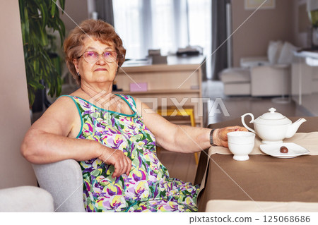 Confident Senior woman sitting indoors, wearing colorful summer dress, enjoying tea time at home with porcelain teapot and chocolate, looking calm and relaxed in cozy modern interior Confident Senior woman sitting indoors, wearing colorful summer dress, enjoying tea time at home with porcelain teapot and chocolate, looking calm and relaxed in cozy modern interior 125068686